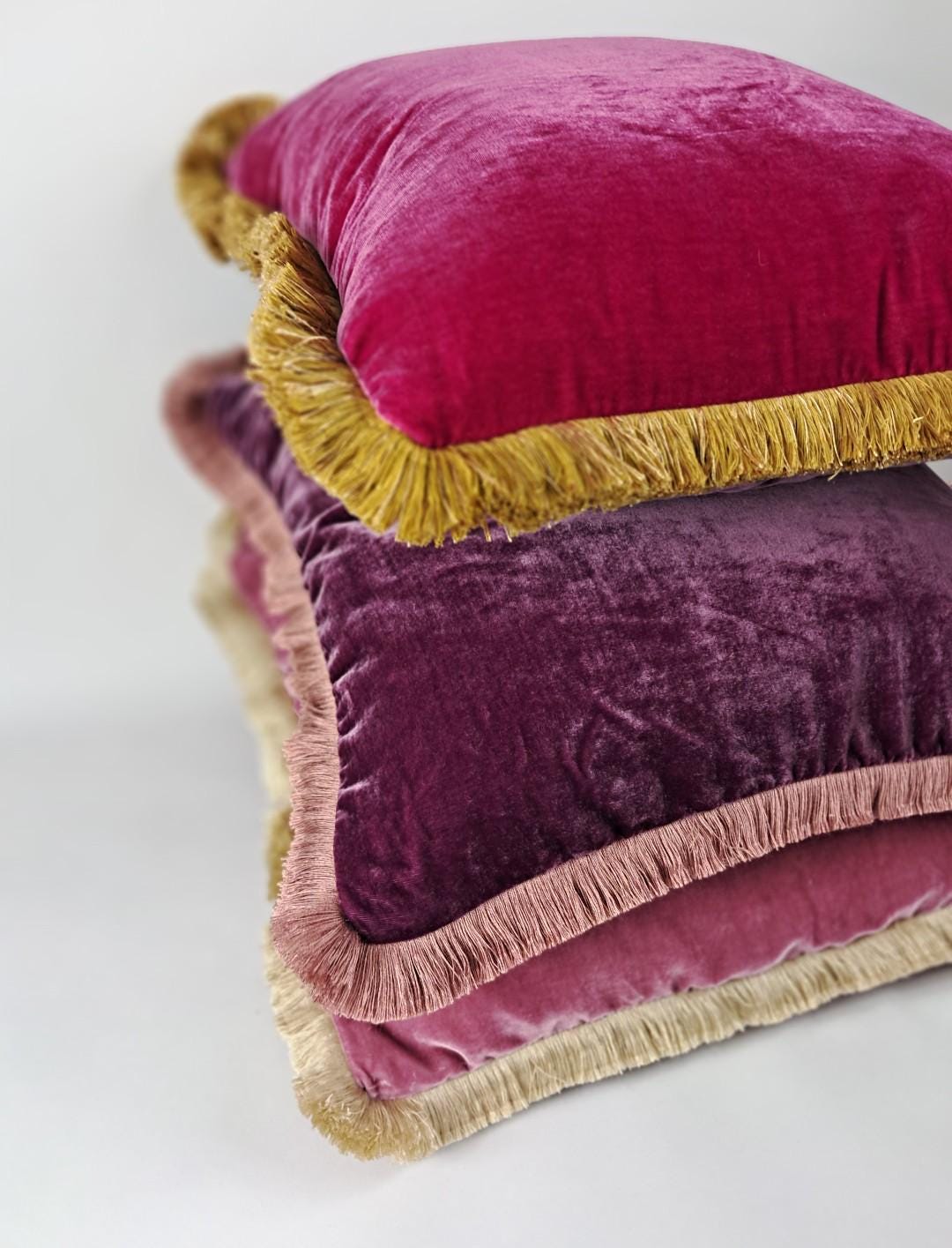 May include: Three velvet cushions in shades of pink and purple, stacked on top of each other. Each square cushion has a contrasting fringe trim. The top cushion is a deep magenta, the middle is a dark purple, and the bottom is a light pink.