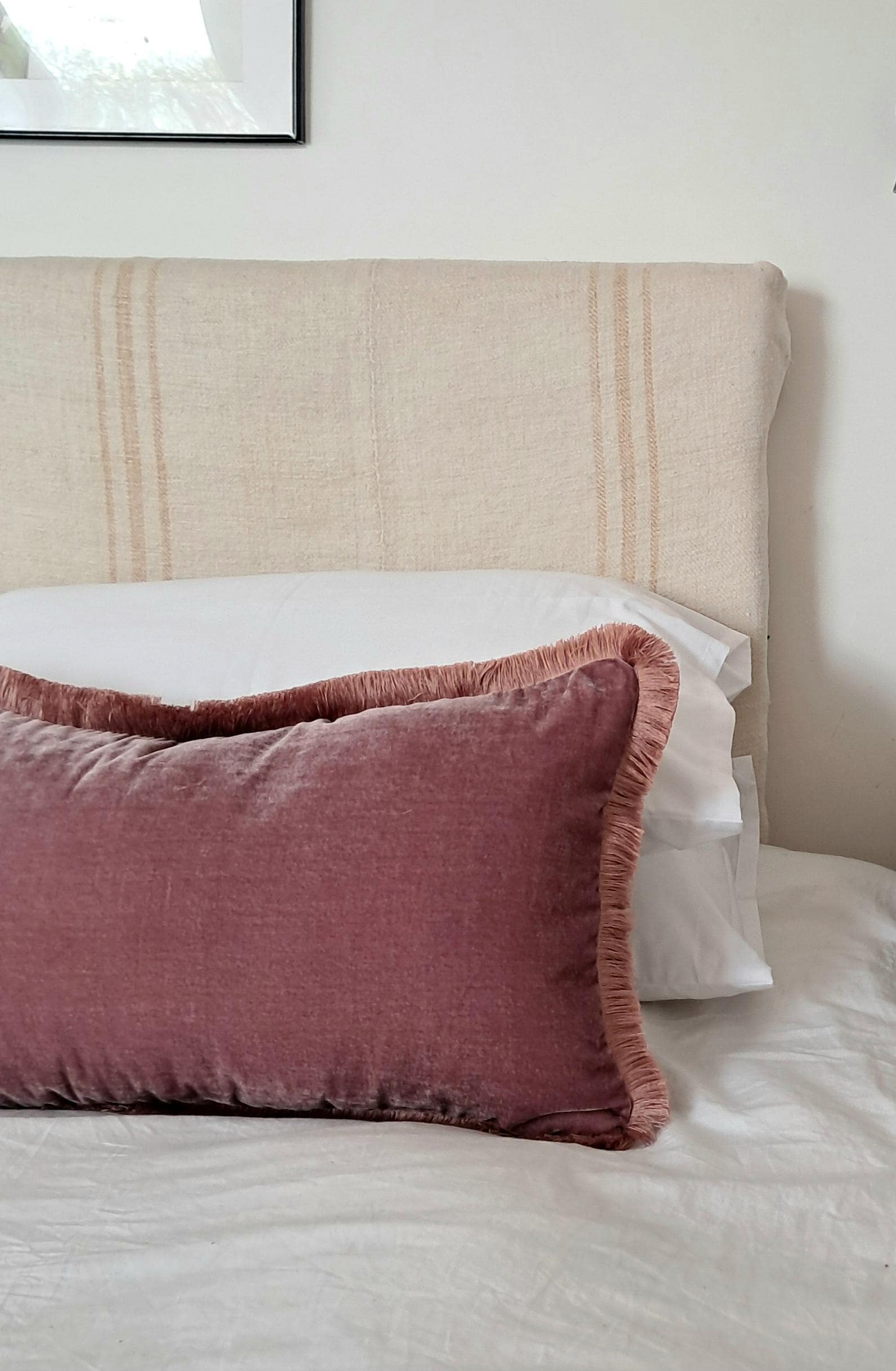 May include: A rectangular, dusty rose velvet cushion with fringing sits on a white bed. The cushion is in front of a white bed sheet and a headboard with vertical stripes.