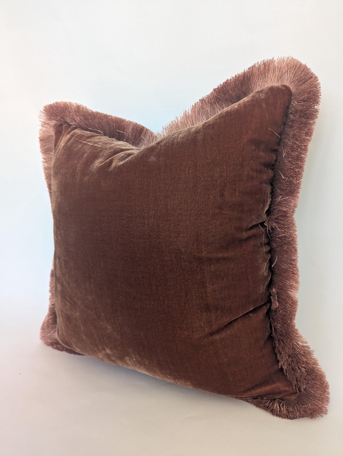 May include: A square, brown velvet cushion with a fringe border. The cushion has a soft, plush appearance and is a warm, earthy tone. The fringe adds texture and a decorative element to the cushion.