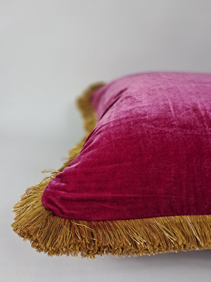 May include: A magenta velvet cushion with a gold fringe trim. The soft texture and rich colour of the cushion create a luxurious aesthetic. The gold fringe adds a decorative element, enhancing the overall design.